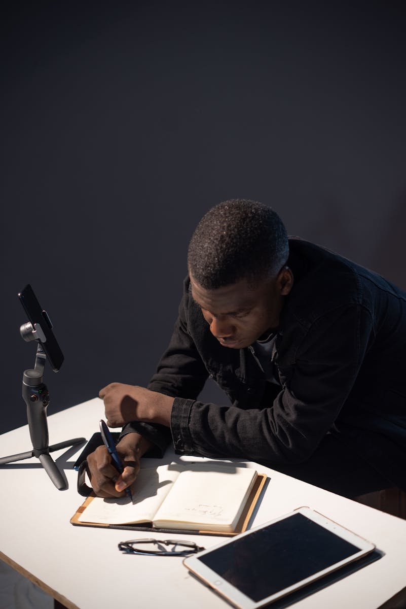 Young man using tech gadgets and writing notes, focusing in a dark room setting.